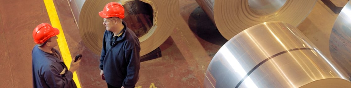 Two factory workers standing next to rolls of steel