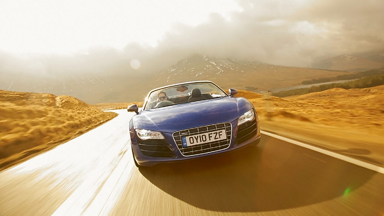  Open top blue car, face on view, driving through the countryside surrounded by mountain scenary