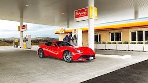 A red Ferrari sitting on a Shell station forecourt with a man leaning on a petrol pump