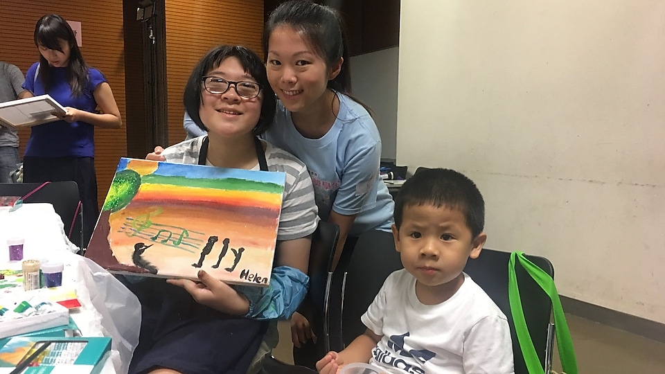 Student with special educational needs held her painting and took photo with parent and volunteer.