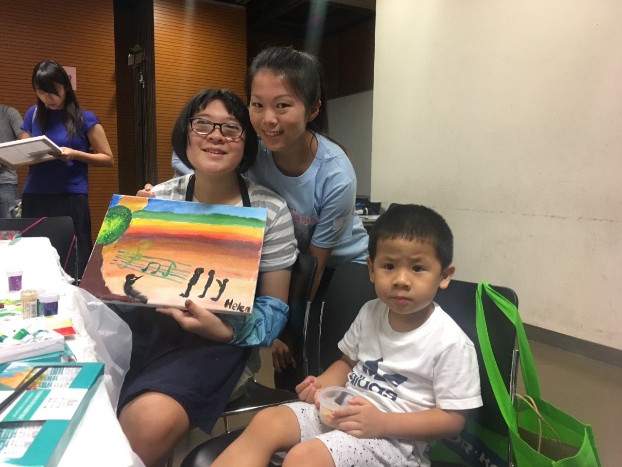 Student with special educational needs held her painting and took photo with parent and volunteer.